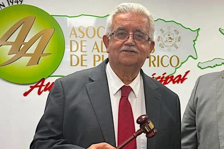 Hombre mayor con traje gris y corbata roja sostiene un mazo frente a un rótulo de la Asociación de Alcaldes de Puerto Rico en una pared blanca.