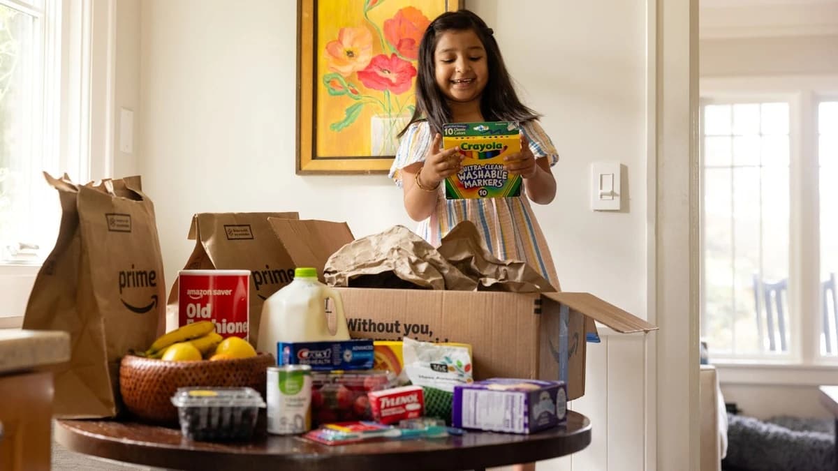 Niña feliz sostiene una caja de marcadores Crayola tras recibir un pedido de Amazon Prime con comestibles y productos esenciales en la mesa de su casa. Imagen suministrada.
