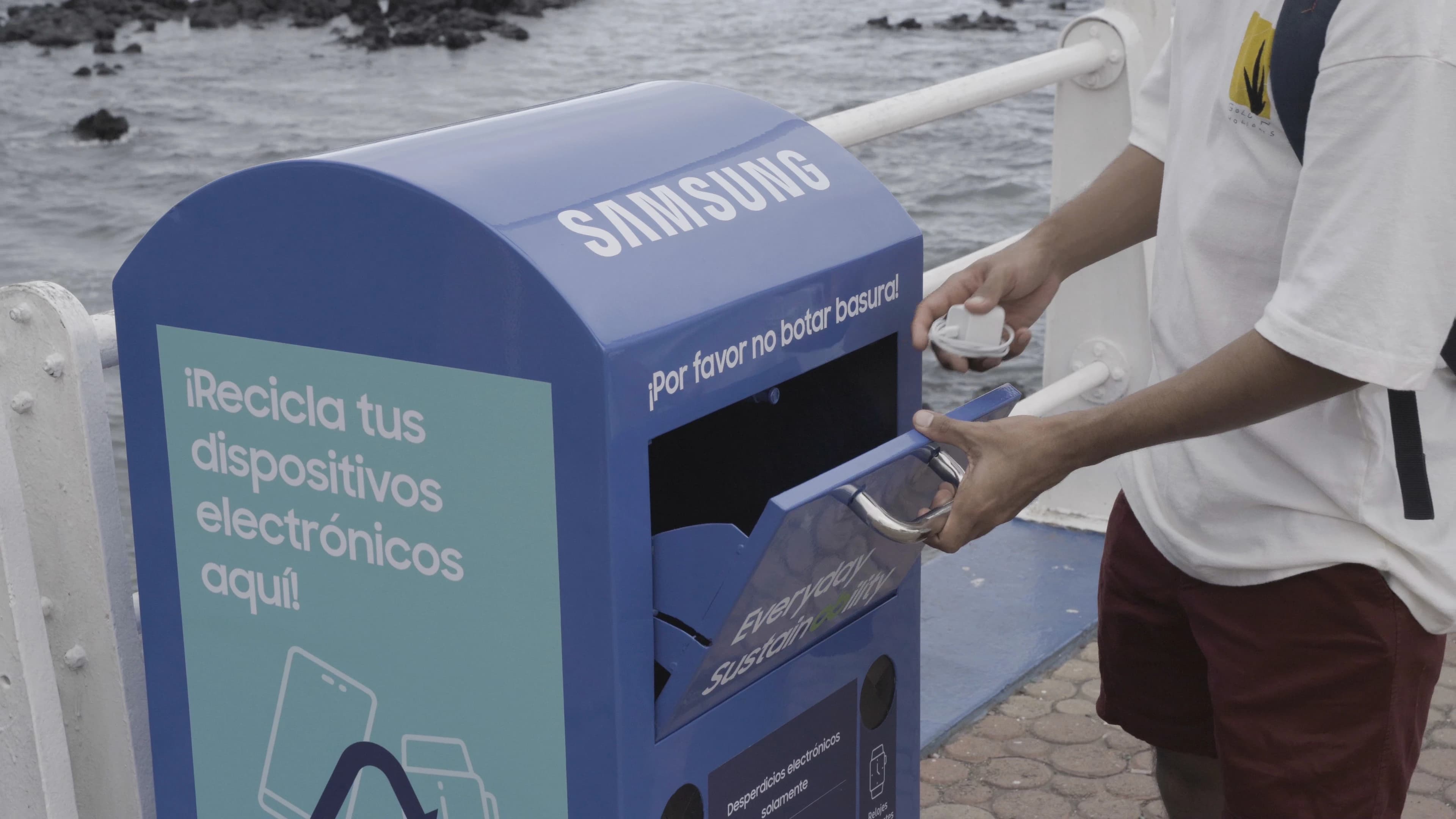 Persona recicla un dispositivo en un contenedor azul de Samsung ubicado junto al mar, diseñado para la recolección responsable de desechos electrónicos.