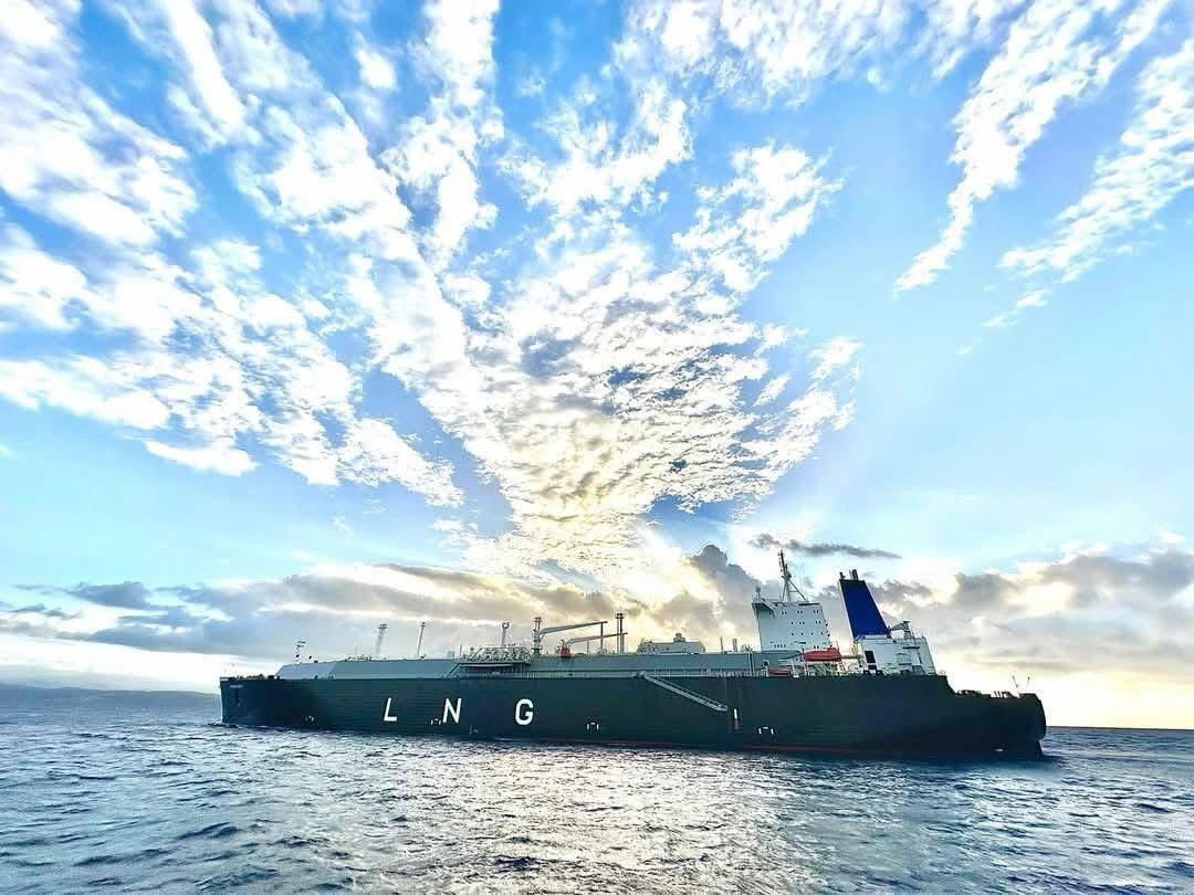 Un buque tanquero de casco oscuro con las letras blancas LNG pintadas en su costado navega en mar abierto, con la superestructura blanca y azul hacia la popa, bajo un cielo dramático de nubes dispersas iluminadas por el sol y una franja de costa apenas visible en el horizonte a la izquierda.