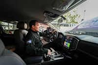 Gustavo Villatoro, vestido de negro con un parche de la bandera de El Salvador en el brazo, sentado al volante de una pickup policial nueva. Con una mano sostiene el guía y con la otra opera los controles de la consola central, junto a una pantalla táctil grande en el tablero. El parabrisas conserva el rótulo invertido de Policía Nacional Civil y el interior mantiene los plásticos protectores de fábrica.