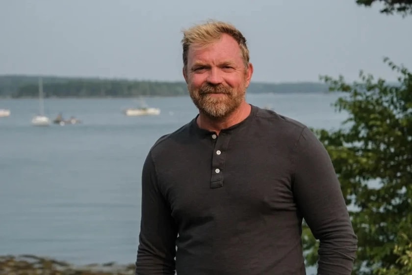 Retrato de Graham Platner, hombre de cabello rubio y barba rojiza, vestido con camisa tipo henley gris oscuro, de pie frente a una bahía con veleros anclados y vegetación a la derecha, en un paisaje costero de Maine.