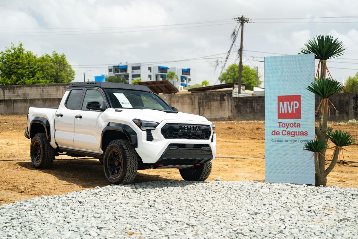 Una pickup Toyota blanca con acentos negros, parrilla con letras TOYOTA y canasta de techo, estacionada sobre terreno de tierra junto a un área de gravilla. A su lado, una pantalla LED vertical muestra el logo de MVP Toyota de Caguas con el lema "Conduce tu mejor jugada", flanqueada por plantas de yuca. Al fondo se ven un muro de hormigón, postes del tendido eléctrico, edificios y un cielo nublado.