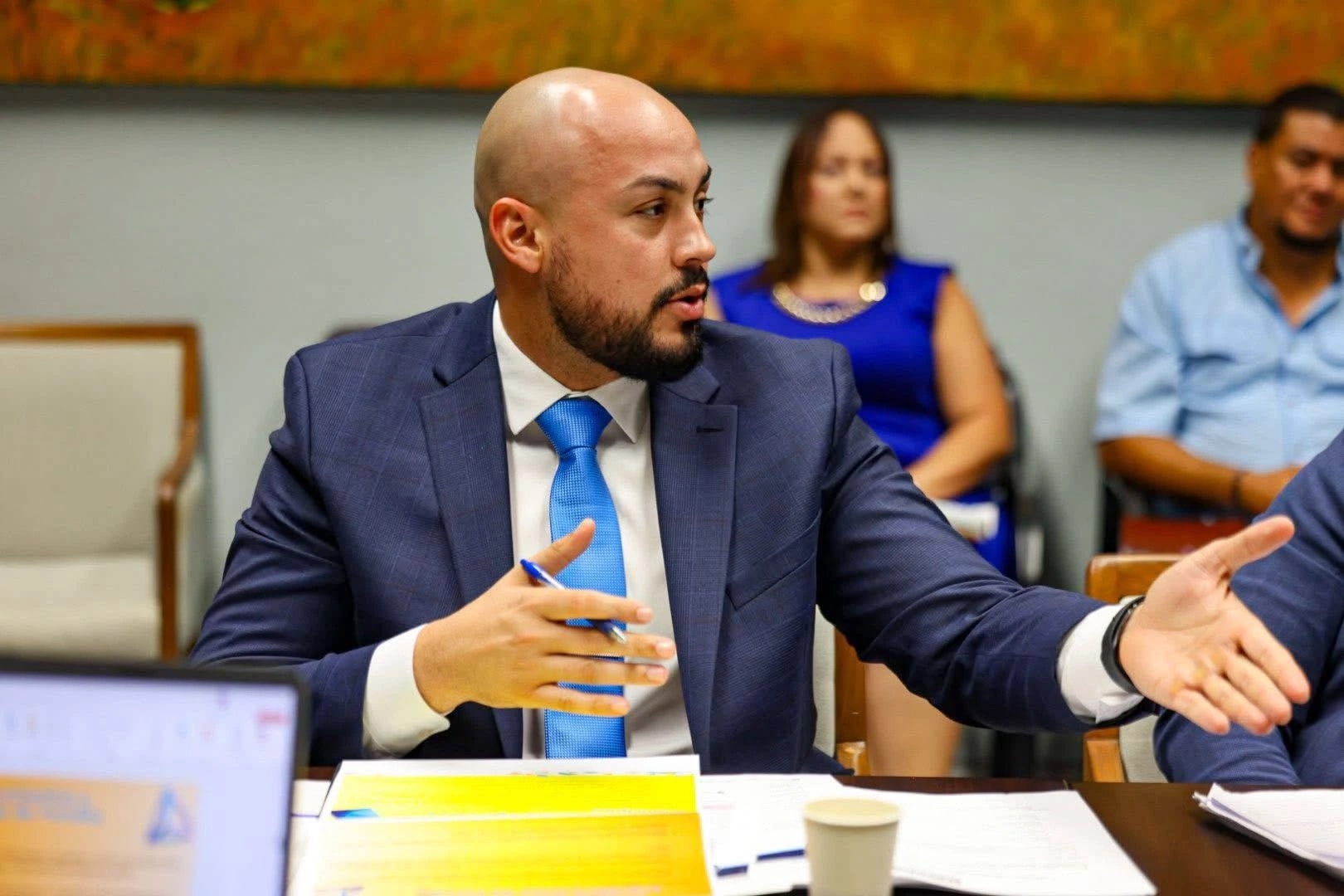 Ángel Morales Vázquez gesticula mientras participa en una reunión de trabajo, vestido con traje azul y corbata azul claro, con documentos sobre la mesa. (Imagen suministrada)