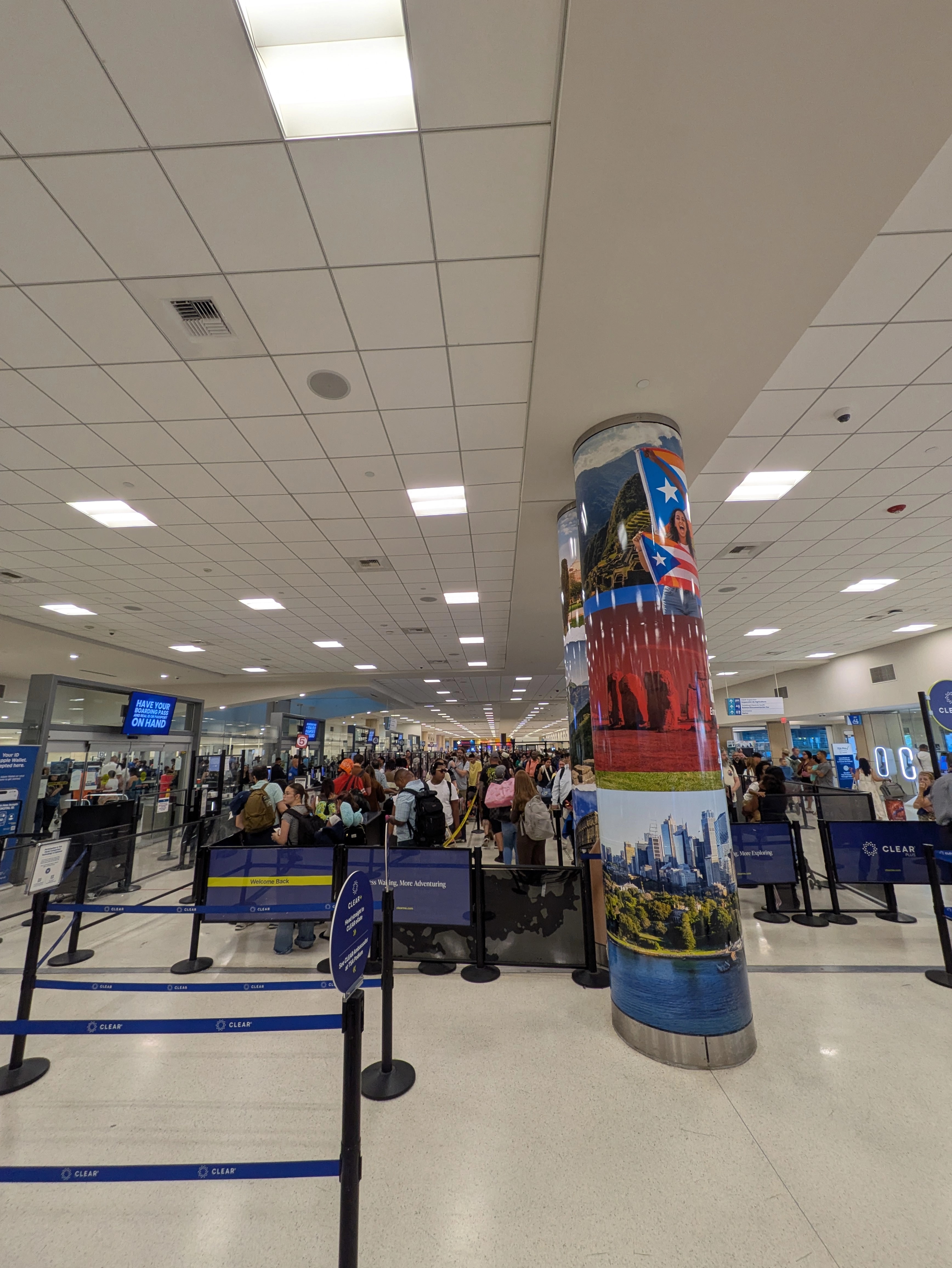 Interior del área de seguridad del Aeropuerto Luis Muñoz Marín. A la derecha, los carriles del servicio CLEAR aparecen vacíos y delimitados con postes y cintas azules, junto a una columna decorada con imágenes de destinos turísticos y una bandera de Puerto Rico. A la izquierda, una larga fila de viajeros con equipaje espera para pasar por los puntos de inspección de la TSA.
