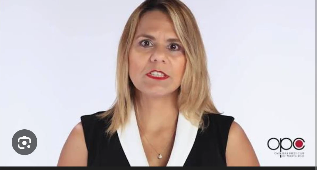 Mujer frente a fondo blanco, usando chaqueta negra y blusa blanca, durante un mensaje institucional del Overseas Press Club de Puerto Rico; se observa el logo del OPC en la esquina inferior derecha.