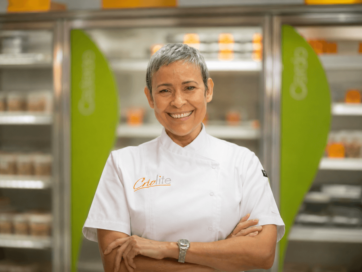 Retrato de la chef Rosa M. García Flores sonriendo con los brazos cruzados, vestida con filipina blanca de chef con el logo de Criolite bordado, frente a neveras comerciales con paneles verdes y productos empacados en un fondo desenfocado.