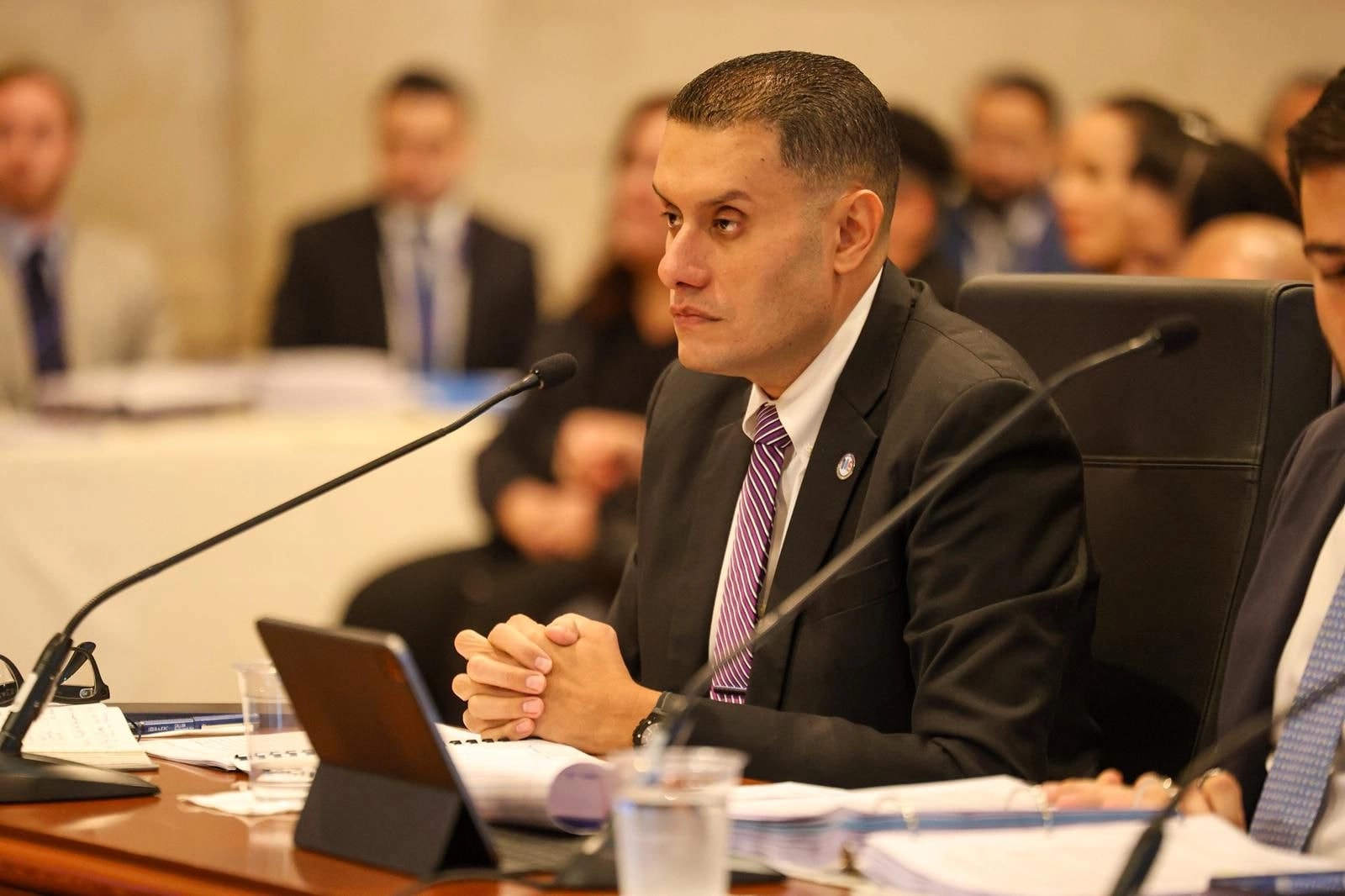 El alcalde de Camuy y presidente de la Federación de Alcaldes, Gabriel Hernández Rodríguez, sentado frente a un micrófono con las manos entrelazadas sobre la mesa durante una vista pública en el Senado de Puerto Rico. Viste traje negro con corbata morada y blanca. Al fondo se observan otros asistentes en la sala.
