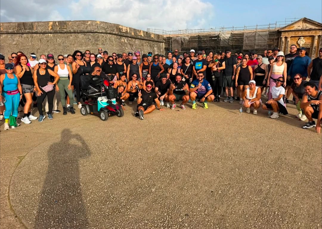 Un grupo numeroso de personas en ropa deportiva posa para una foto grupal frente a una muralla colonial de piedra bajo la luz de la mañana. Entre los participantes hay una persona en un scooter motorizado de movilidad. A la derecha se aprecia una estructura neoclásica con columnas cubierta parcialmente por andamios. En el primer plano, la sombra del fotógrafo se proyecta sobre el pavimento.