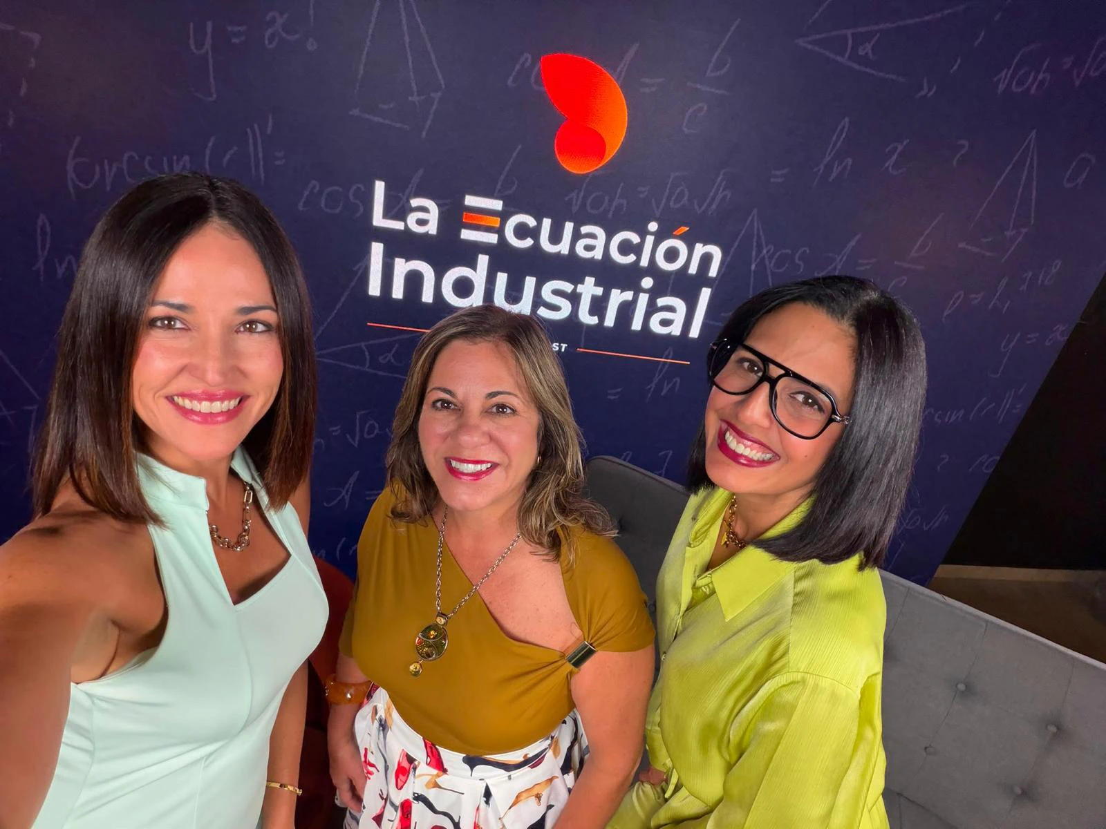 Tres mujeres sonrientes posan en el estudio del pódcast La Ecuación Industrial, frente a una pared azul oscuro decorada con fórmulas matemáticas y el logotipo del programa con un ícono naranja. A la izquierda, una mujer de cabello castaño con blusa verde menta; al centro, una mujer de blusa mostaza y falda estampada; a la derecha, una mujer de espejuelos oscuros y camisa verde lima, junto a un sofá gris.