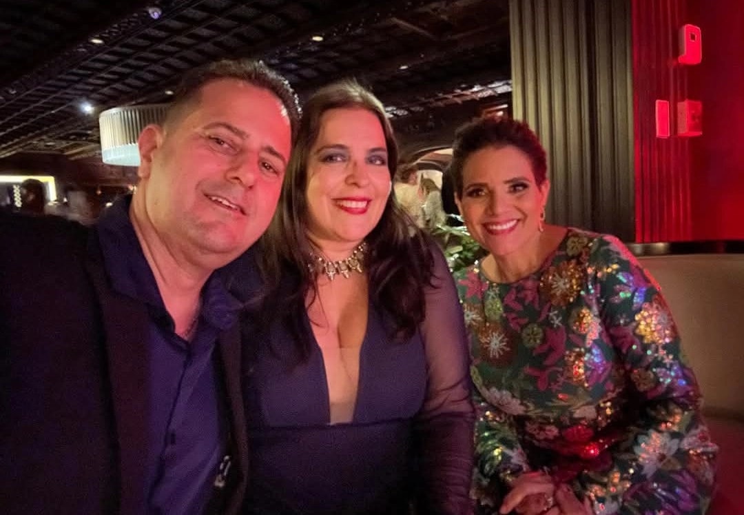 Fotografía tomada en un evento nocturno en la que aparecen tres personas posando juntas. A la izquierda, el secretario de Corrección Francisco Quiñones Rivera con camisa oscura. Al centro, Lisoannette González Ruiz con vestido azul marino y collar. A la derecha, Janet Parra con vestido floreado multicolor. Al fondo se observa iluminación tenue y cortinas rojas.