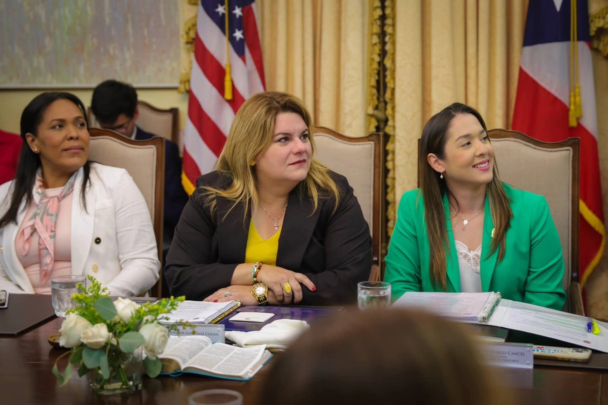 Tres mujeres sentadas a una mesa de conferencias de madera pulida en un salón formal con cortinas doradas y las banderas de Estados Unidos y de Puerto Rico al fondo. A la izquierda, la secretaria de Estado, Rosachely Rivera Santana, de cabello oscuro y blazer blanco sobre blusa rosada con estampado, mira hacia un lado. En el centro, la gobernadora Jenniffer González Colón, rubia, con blazer oscuro sobre blusa amarilla, mantiene las manos entrelazadas sobre la mesa. A la derecha, la directora de Turismo, Willianette Robles Cancel, de cabello oscuro y blazer verde sobre blusa blanca de encaje, sonríe. Sobre la mesa hay un libro abierto, carpetas con documentos, vasos de agua, placas con nombres y un arreglo de rosas blancas; en primer plano se ve la parte posterior de la cabeza de otra persona.