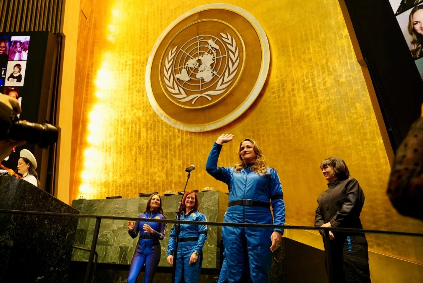 Deborah Martorell, vestida con un traje espacial azul de Blue Origin, saluda con la mano en el podio del Salón de la Asamblea General de las Naciones Unidas en Nueva York. Al fondo se aprecia el emblema dorado de la ONU. Otras mujeres también con trajes azules acompañan en el escenario.