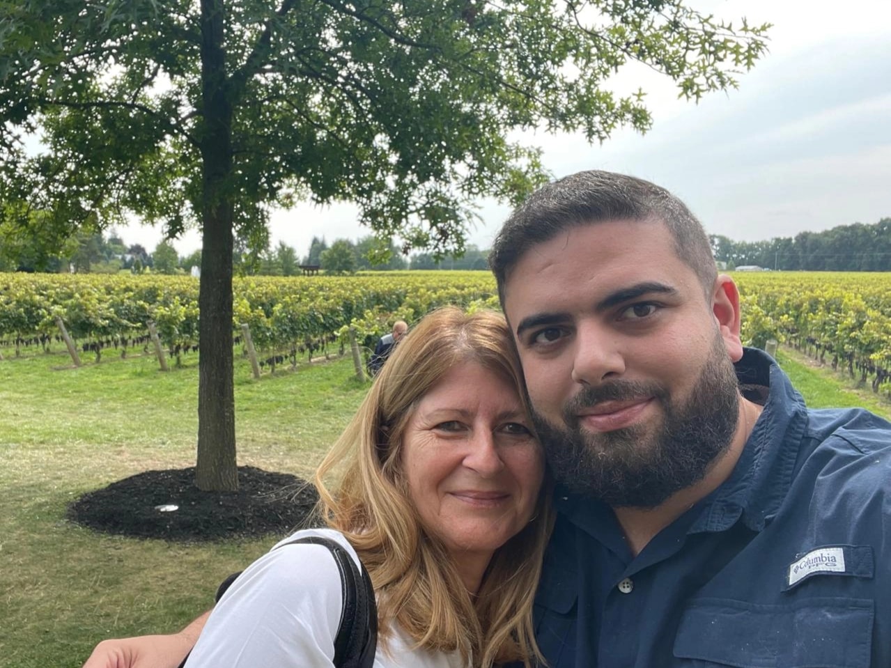 A selfie of a smiling middle-aged woman with blonde hair and a young man with a dark beard, posing together outdoors. They are standing in front of a sprawling green vineyard under a large leafy tree. The man is wearing a blue Columbia button-down shirt.