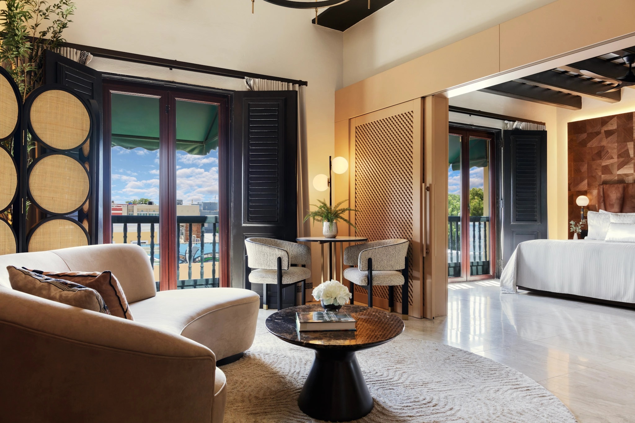 Interior de la suite presidencial del Hotel El Convento mostrando una sala con sofá beige, mesa de mármol oscuro, sillas de diseño contemporáneo, balcones coloniales con persianas negras y toldos verdes abiertos hacia el Viejo San Juan, techos altos con vigas de madera oscura, y la habitación con cama al fondo.