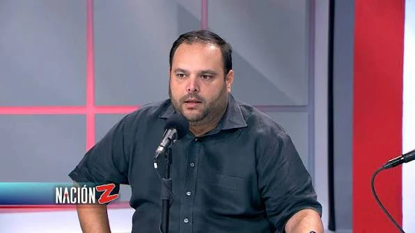 Hombre de cabello oscuro y barba corta, con camisa negra de botones, sentado frente a un micrófono en un estudio de televisión con paneles rojos y grises al fondo. En la esquina inferior izquierda aparece el logo del programa Nación Z.