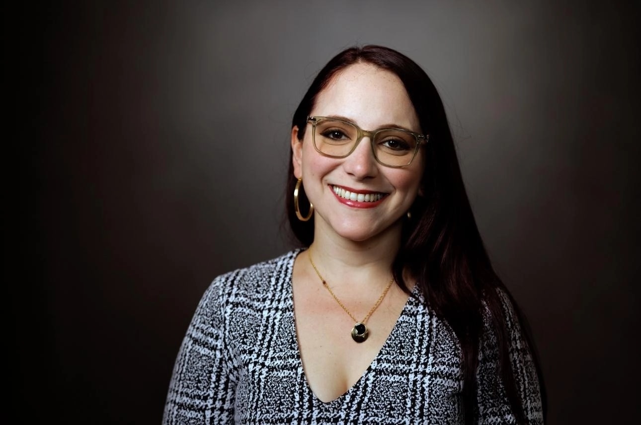 Retrato profesional de la licenciada Claudia Méndez. Mujer de cabello largo castaño rojizo, espejuelos de montura rectangular, aretes de argolla dorados y collar con dije. Viste una blusa de cuadros en blanco y negro. Sonríe frente a un fondo gris oscuro.