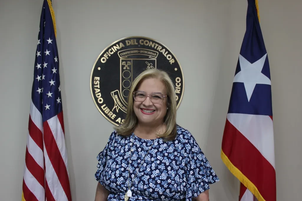 Yesmín Valdivieso, contralora de Puerto Rico, posa frente al emblema oficial de la Oficina del Contralor junto a las banderas de Puerto Rico y Estados Unidos.