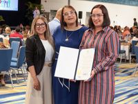 Tres mujeres sonrientes posan de pie en un salón de actividades con alfombra azul de patrones ondulados y mesas con manteles dorados al fondo. La de la izquierda viste chaqueta negra sobre blusa blanca y falda beige; la del centro, un vestido azul con collar dorado y espejuelos claros; y la de la derecha, un vestido de rayas anaranjadas y azules. Las dos últimas sostienen juntas una carpeta azul abierta con una proclama oficial firmada, con el sello dorado visible.