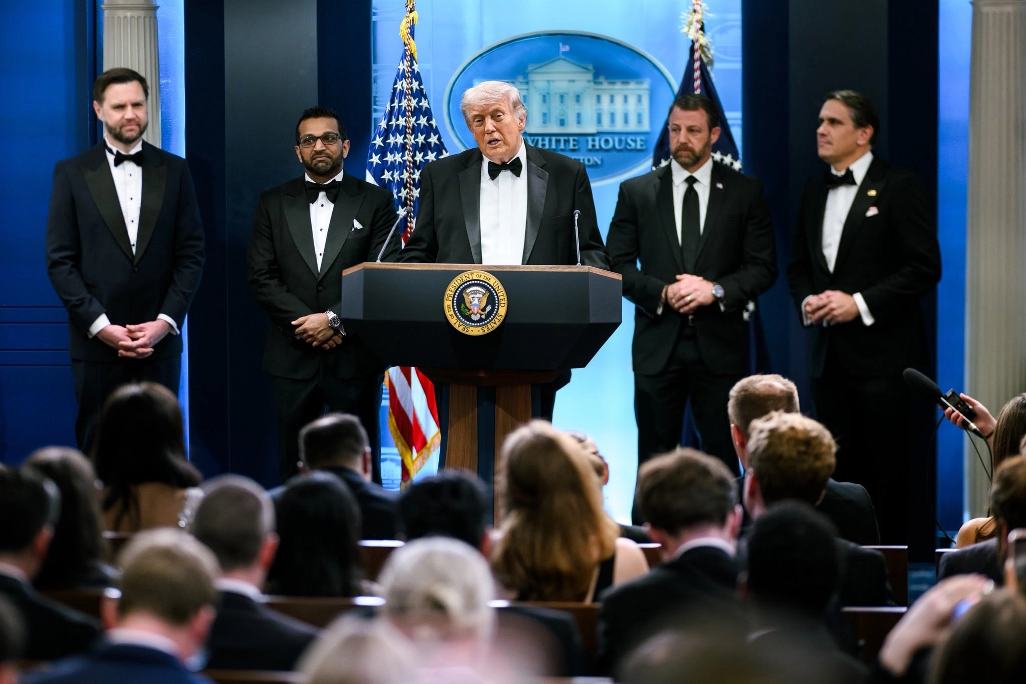 El presidente Donald Trump habla desde un podio con el sello presidencial en la Sala de Briefing de la Casa Blanca, vestido de esmoquin negro. A su izquierda se encuentran el vicepresidente JD Vance y otros funcionarios, también de esmoquin. Al fondo se observan banderas estadounidenses y el logo de la Casa Blanca. Frente al podio se aprecia una audiencia sentada.