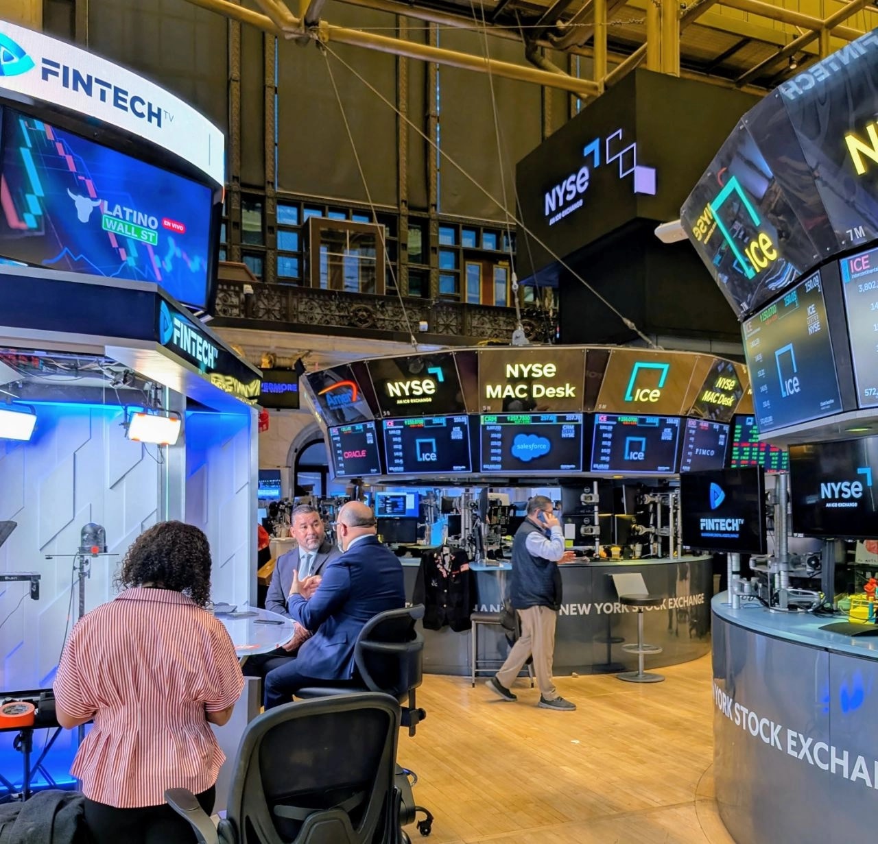 Interior del piso de operaciones del New York Stock Exchange con pantallas de cotizaciones, el escritorio MAC Desk del NYSE y personas conversando frente al set de Latino Wall St. de FintechTV.