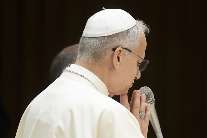 El papa León XIV, vestido de blanco y con solideo, ora con las manos juntas mientras sostiene un micrófono durante una audiencia general en el Aula Pablo VI del Vaticano.