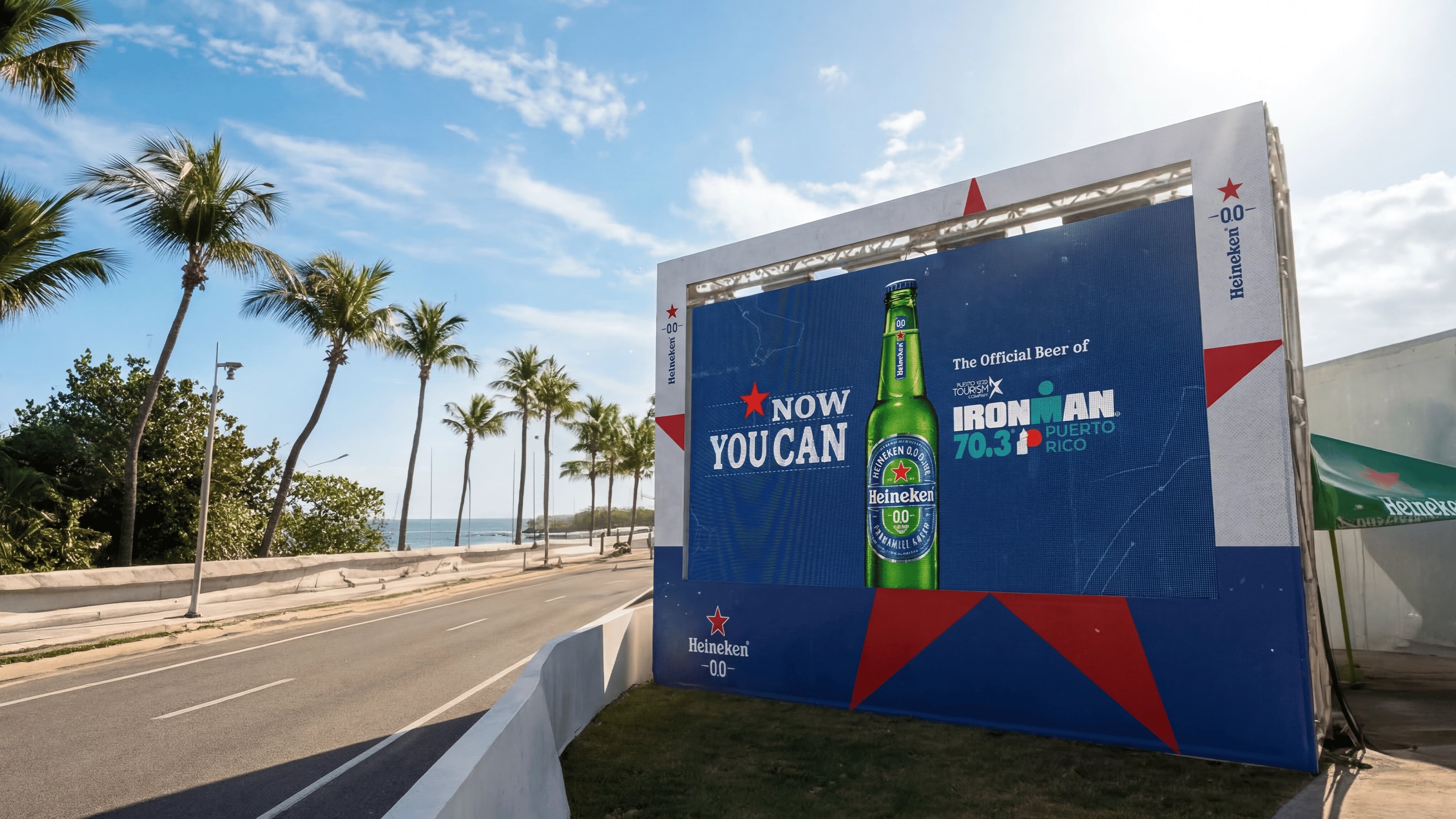 Pantalla gigante de Heineken 0.0 junto al recorrido costero del IRONMAN 70.3 Puerto Rico con el mensaje "Now You Can" y una botella de Heineken 0.0, identificada como la cerveza oficial del evento, con palmeras y el océano al fondo.