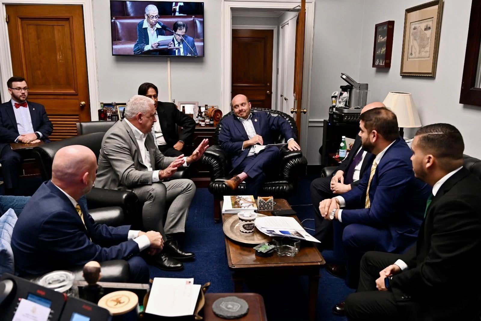 Grupo de seis hombres reunidos en una oficina congresional en Washington, sentados en sofás de cuero negro alrededor de una mesa de centro con documentos, mientras un televisor transmite una sesión legislativa al fondo.