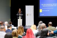 María L. Marín Colón habla desde un podio de acrílico en un salón de conferencias, junto a un banner blanco con el logo de COR3 y frente a una audiencia de funcionarios sentados de espaldas a la cámara, con una pantalla azul al fondo que proyecta una presentación sobre el uso de subastas en proyectos subvencionados con fondos federales.