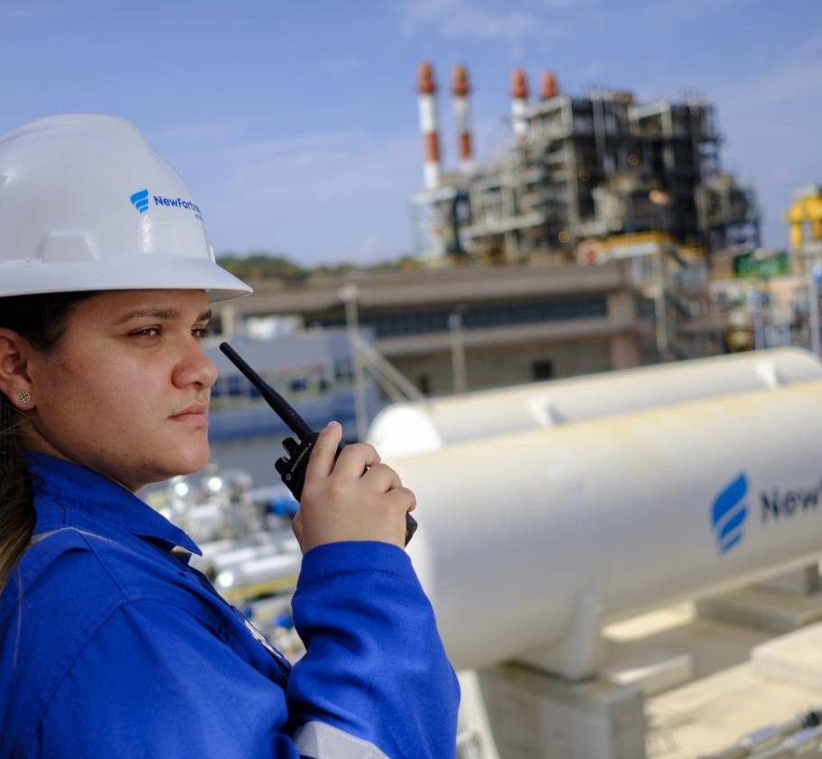 Empleada de New Fortress Energy con casco blanco y uniforme azul sostiene un radio de comunicación frente a una planta de generación eléctrica con tanques y chimeneas al fondo.