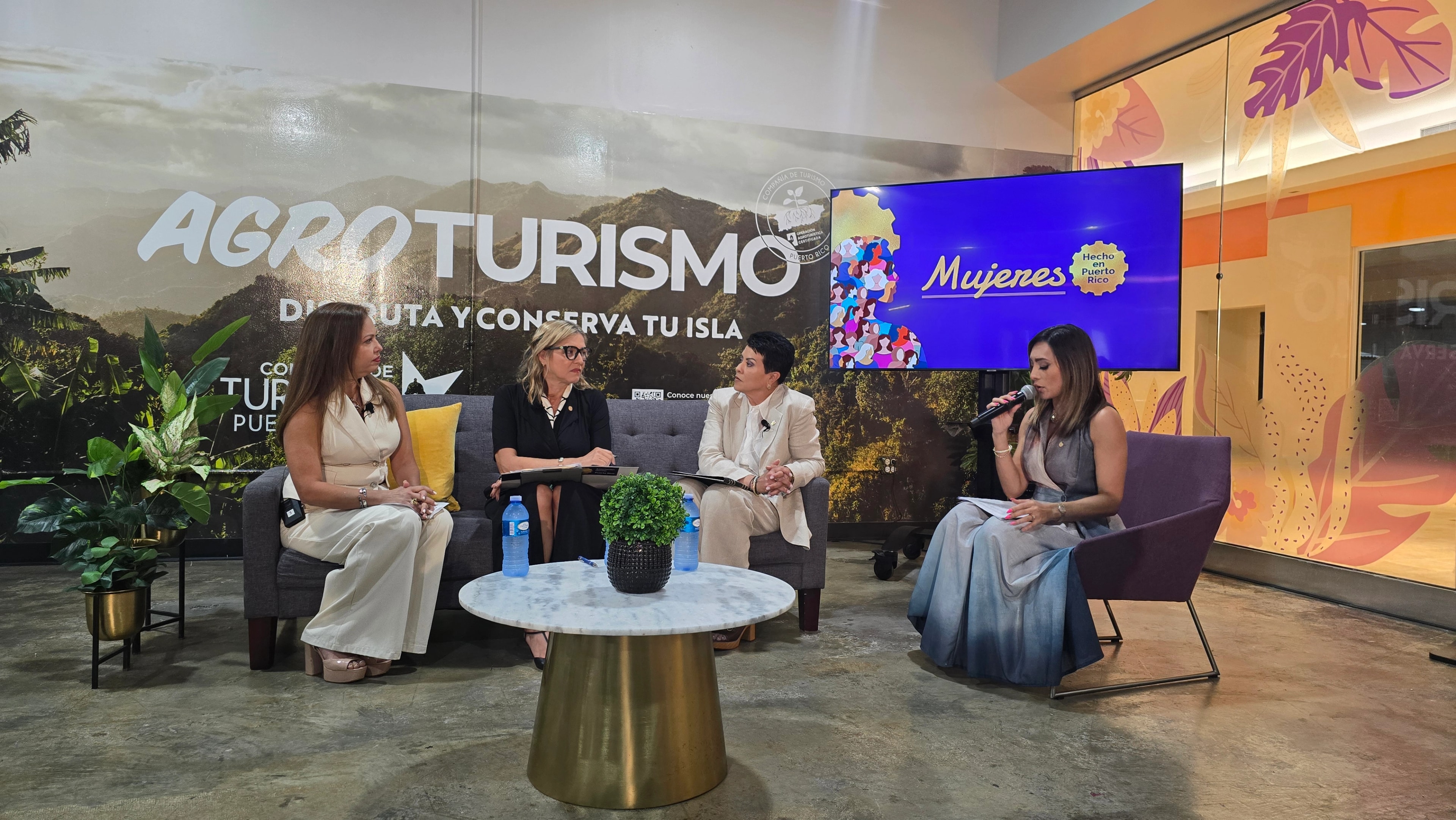 Cuatro mujeres participan en un conversatorio en panel. Tres de ellas están sentadas en un sofá gris y una, con micrófono en mano, modera desde una silla separada a la derecha. Al fondo se ve una pantalla con el logo de "Mujeres Hecho en Puerto Rico" y un mural de Agroturismo Puerto Rico.