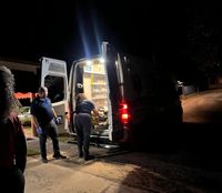 Escena nocturna de una ambulancia con las puertas traseras abiertas e iluminada por dentro, mientras dos personas con guantes atienden una camilla en la parte posterior del vehículo, estacionado frente a una residencia.