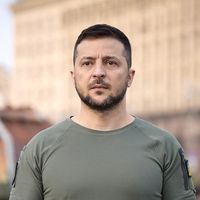 Retrato de Volodymyr Zelensky, de cabello oscuro corto y barba, con expresión seria y mirada al frente, vestido con una camiseta militar verde oliva con un parche de la bandera de Ucrania en la manga, frente a un edificio de fachada clara desenfocado al fondo.