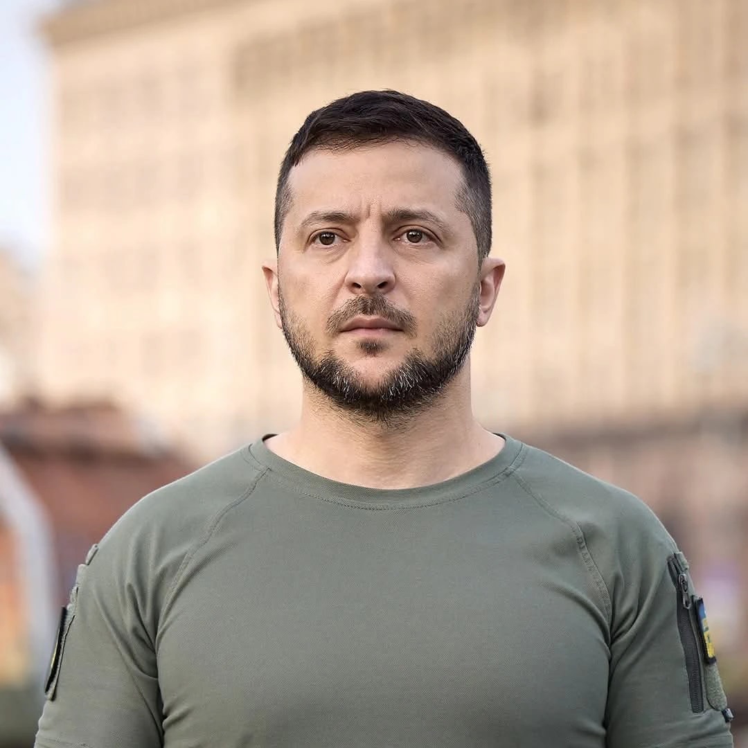 Retrato de Volodymyr Zelensky, de cabello oscuro corto y barba, con expresión seria y mirada al frente, vestido con una camiseta militar verde oliva con un parche de la bandera de Ucrania en la manga, frente a un edificio de fachada clara desenfocado al fondo.