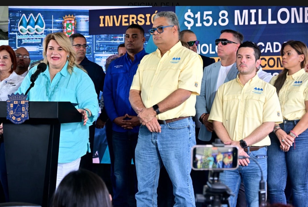 La gobernadora Jenniffer González habla desde un podio durante un evento al aire libre junto a funcionarios de la Autoridad de Acueductos y Alcantarillados vestidos con camisas amarillas, con una pantalla de fondo que muestra una inversión de $15.8 millones