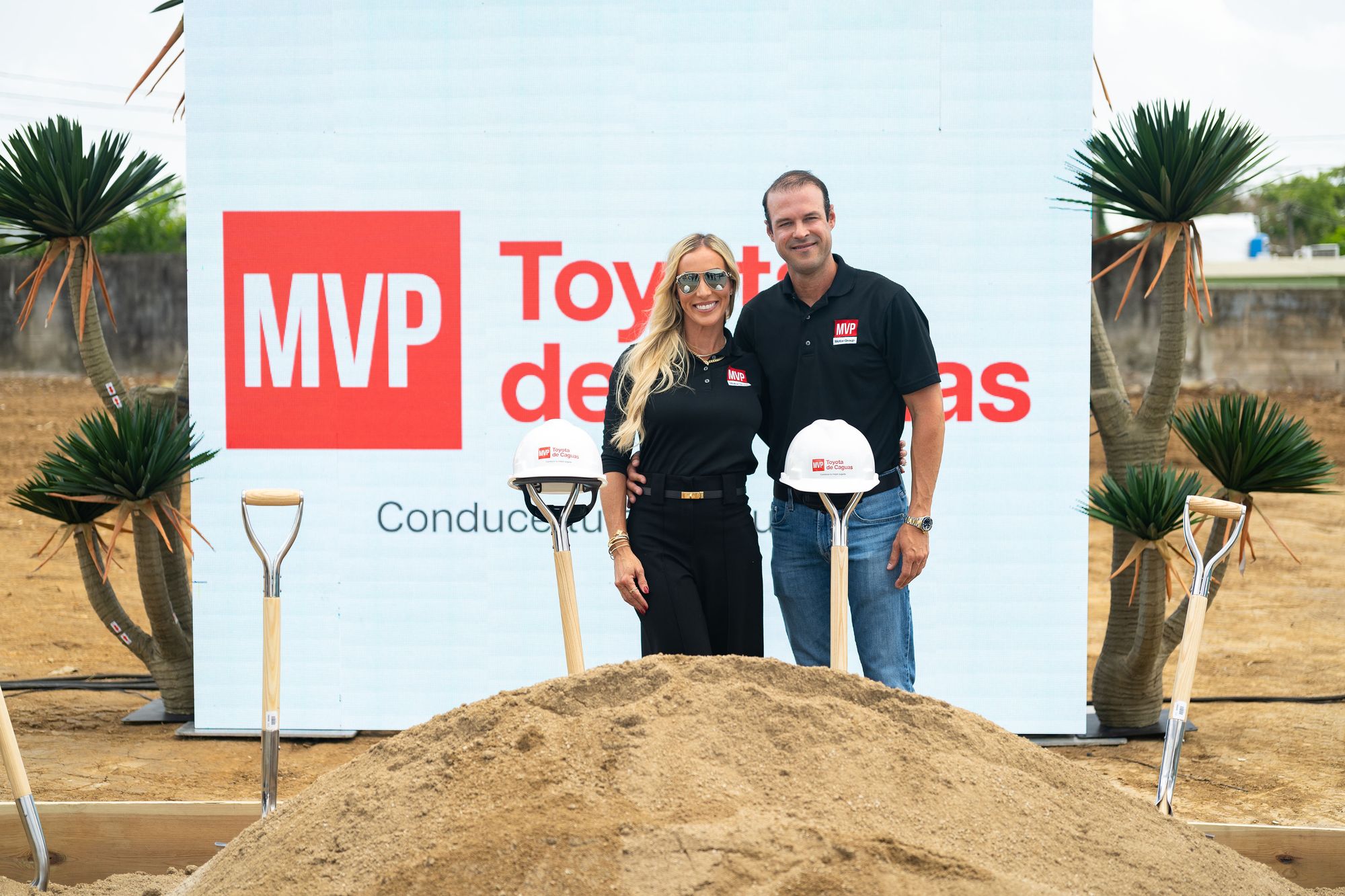 Un hombre y una mujer vestidos con camisas polo negras con el logo de MVP posan sonrientes frente a una pantalla con el logo de MVP Toyota de Caguas, en un terreno en preparación. Frente a ellos hay un montículo de tierra y palas ceremoniales con mangos de madera, dos de ellas con cascos de construcción blancos rotulados con el logo del concesionario. A los lados se ven plantas de yuca y un cielo nublado.