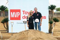 Un hombre y una mujer vestidos con camisas polo negras con el logo de MVP posan sonrientes frente a una pantalla con el logo de MVP Toyota de Caguas, en un terreno en preparación. Frente a ellos hay un montículo de tierra y palas ceremoniales con mangos de madera, dos de ellas con cascos de construcción blancos rotulados con el logo del concesionario. A los lados se ven plantas de yuca y un cielo nublado.