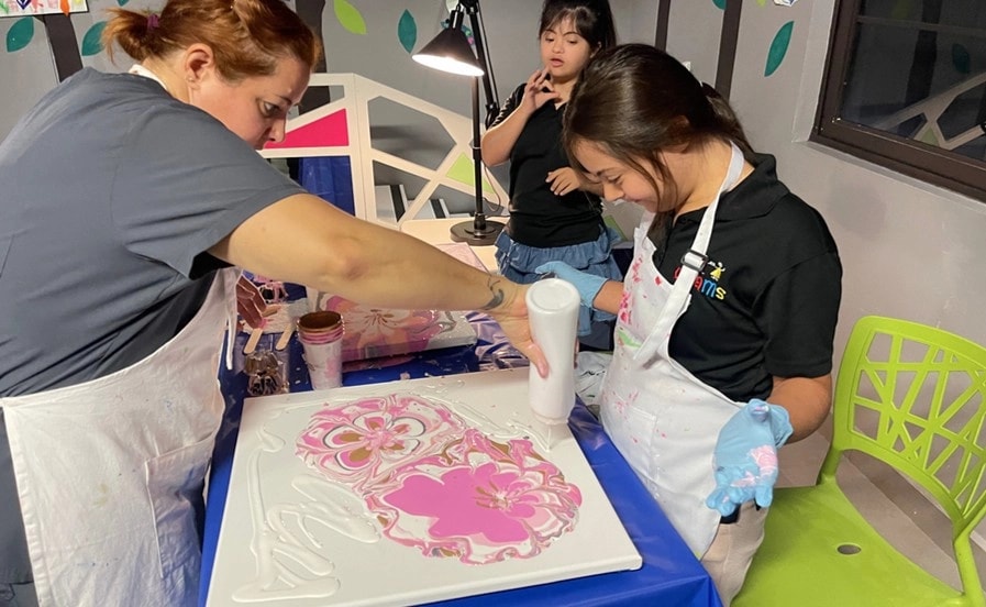 Niña participa en una clase de arte junto a una instructora, aplicando pintura sobre un lienzo en un taller creativo inclusivo organizado por Lily’s Angels, mientras otras participantes observan el proceso.