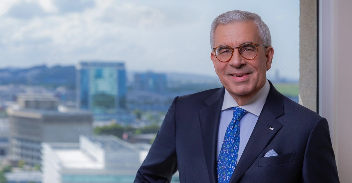 Arturo García-Sola, director general de McConnell Valdés LLC, posa sonriente frente a una ventana con vista panorámica al área metropolitana de San Juan. Viste traje azul oscuro con corbata azul de motivos florales y porta el pin de la firma en la solapa.
