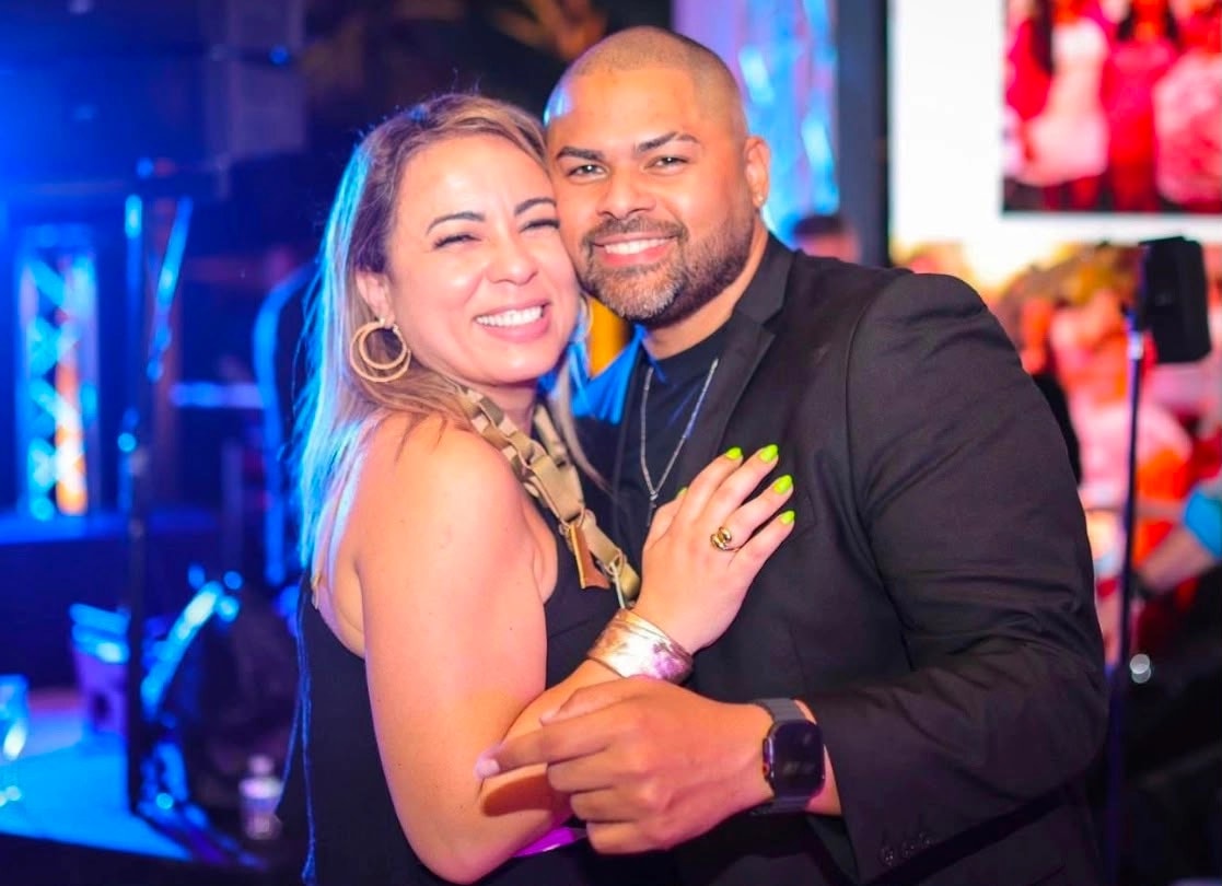 Itza García y Luis Sánchez posan abrazados y sonrientes en un ambiente festivo con iluminación azul y rosada. Ella, de cabello rubio, viste top negro y aretes de aros dorados, con una mano sobre el pecho de él; él, de cabeza rapada y barba, viste chaqueta y camisa negras con un reloj en la muñeca.