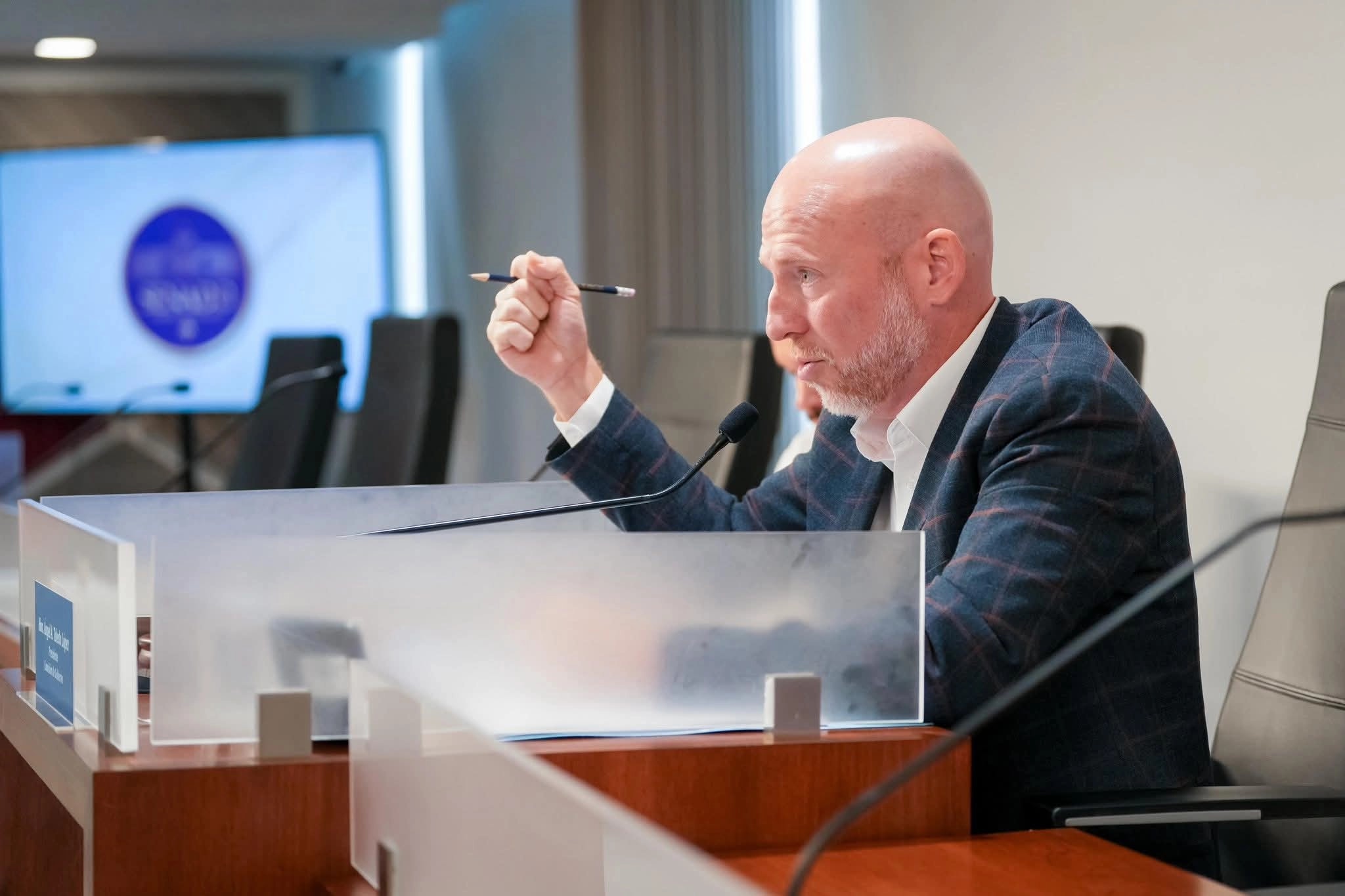 El senador Ángel Toledo sostiene un bolígrafo mientras habla desde su asiento en la sala de la Comisión de Gobierno del Senado, con una pantalla y sillas vacías al fondo.