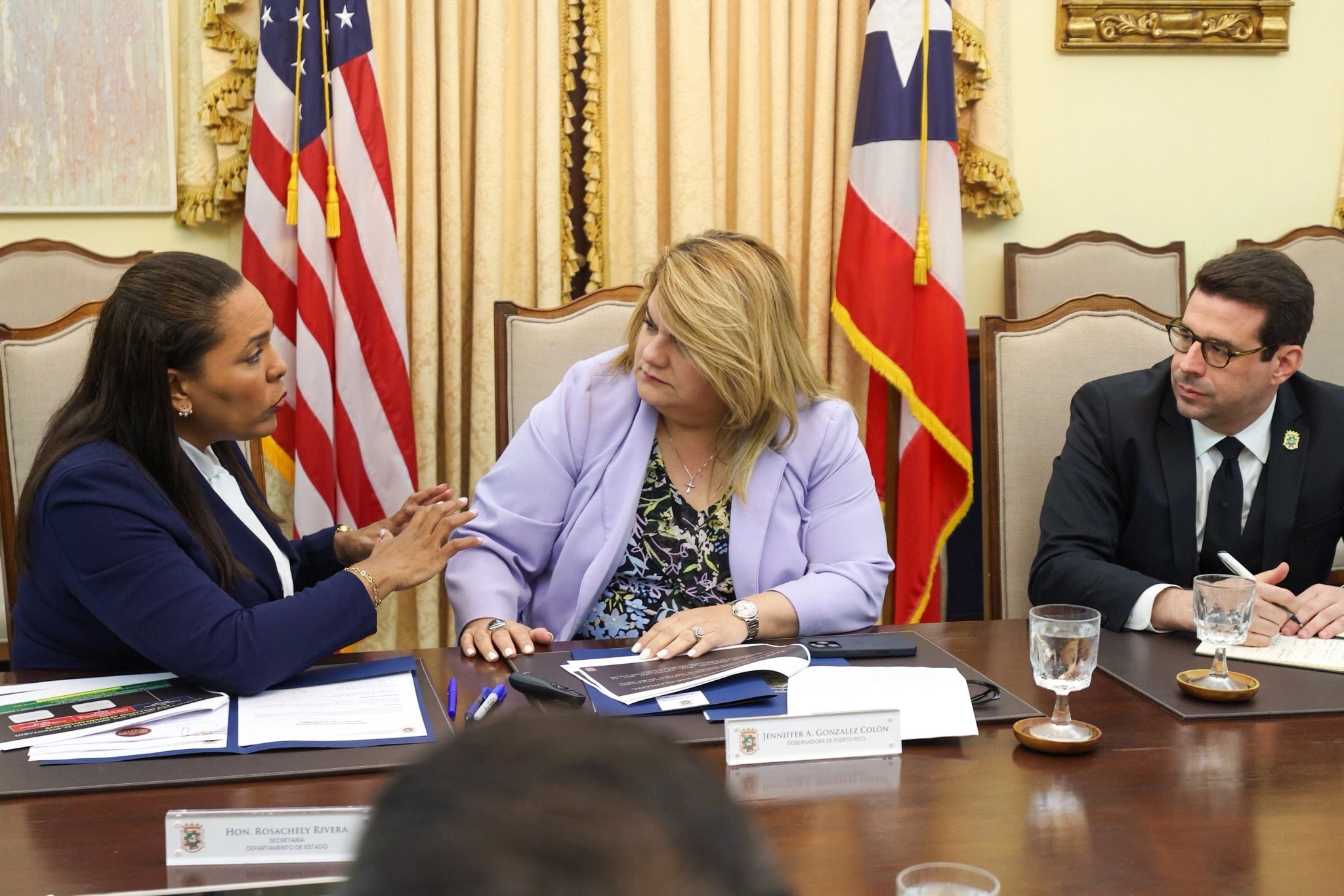 La secretaria de Estado Rosachely Rivera Santana, a la izquierda, conversa con la gobernadora Jenniffer González Colón, al centro, durante una reunión en La Fortaleza. En la mesa se observan las placas identificadoras de ambas funcionarias. A la derecha, un funcionario no identificado toma notas. Al fondo se aprecian las banderas de Estados Unidos y Puerto Rico.