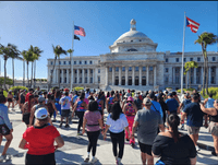 Un grupo numeroso de personas con ropa deportiva, vistas de espaldas, hace ejercicios de pie sobre la explanada de mármol frente al Capitolio de Puerto Rico, con su cúpula blanca al fondo bajo un cielo despejado. A la izquierda ondean la bandera de Estados Unidos y la de los prisioneros de guerra, a la derecha la bandera de Puerto Rico, y palmas reales bordean el edificio.
