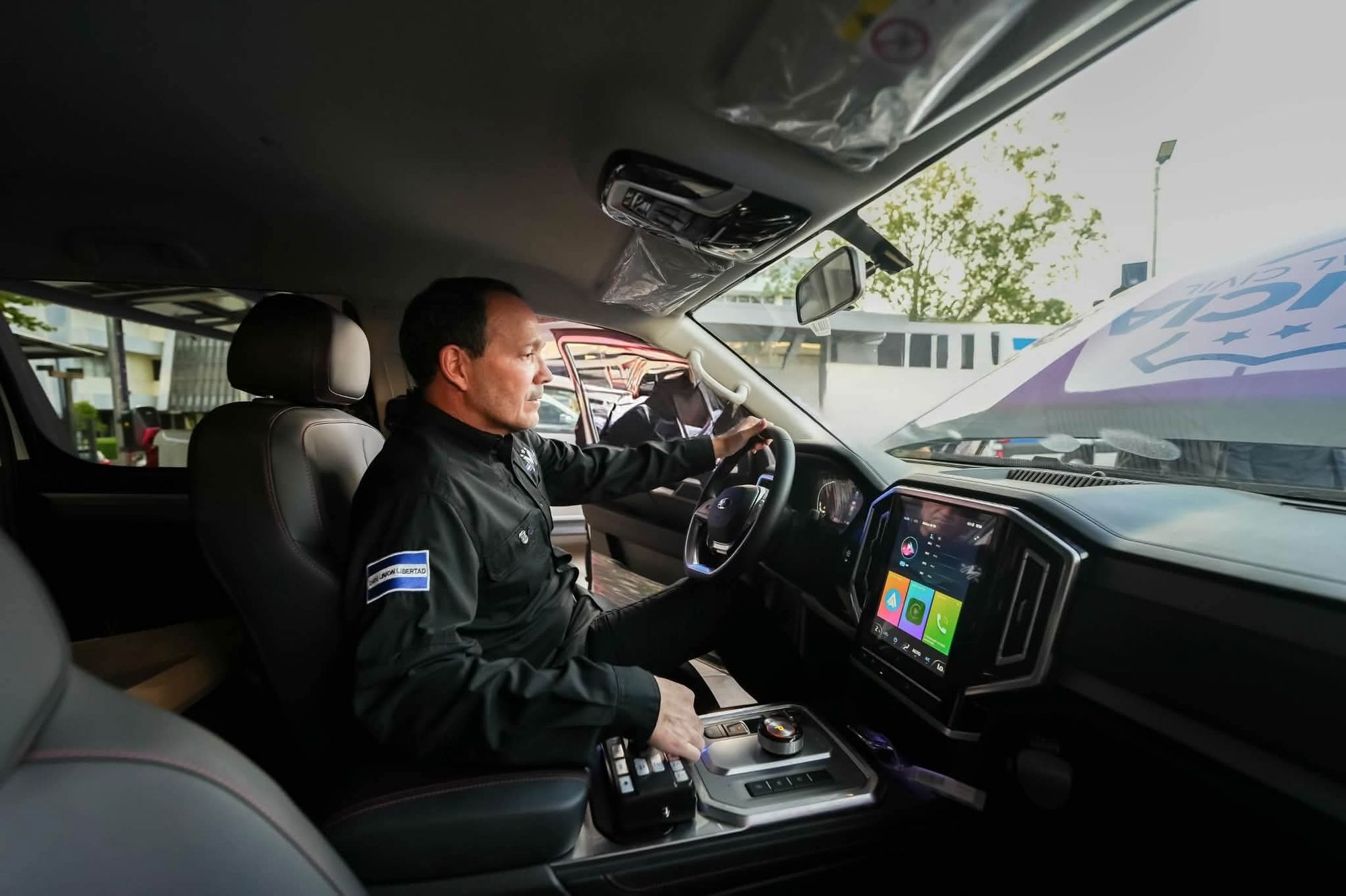 Gustavo Villatoro, vestido de negro con un parche de la bandera de El Salvador en el brazo, sentado al volante de una pickup policial nueva. Con una mano sostiene el guía y con la otra opera los controles de la consola central, junto a una pantalla táctil grande en el tablero. El parabrisas conserva el rótulo invertido de Policía Nacional Civil y el interior mantiene los plásticos protectores de fábrica.