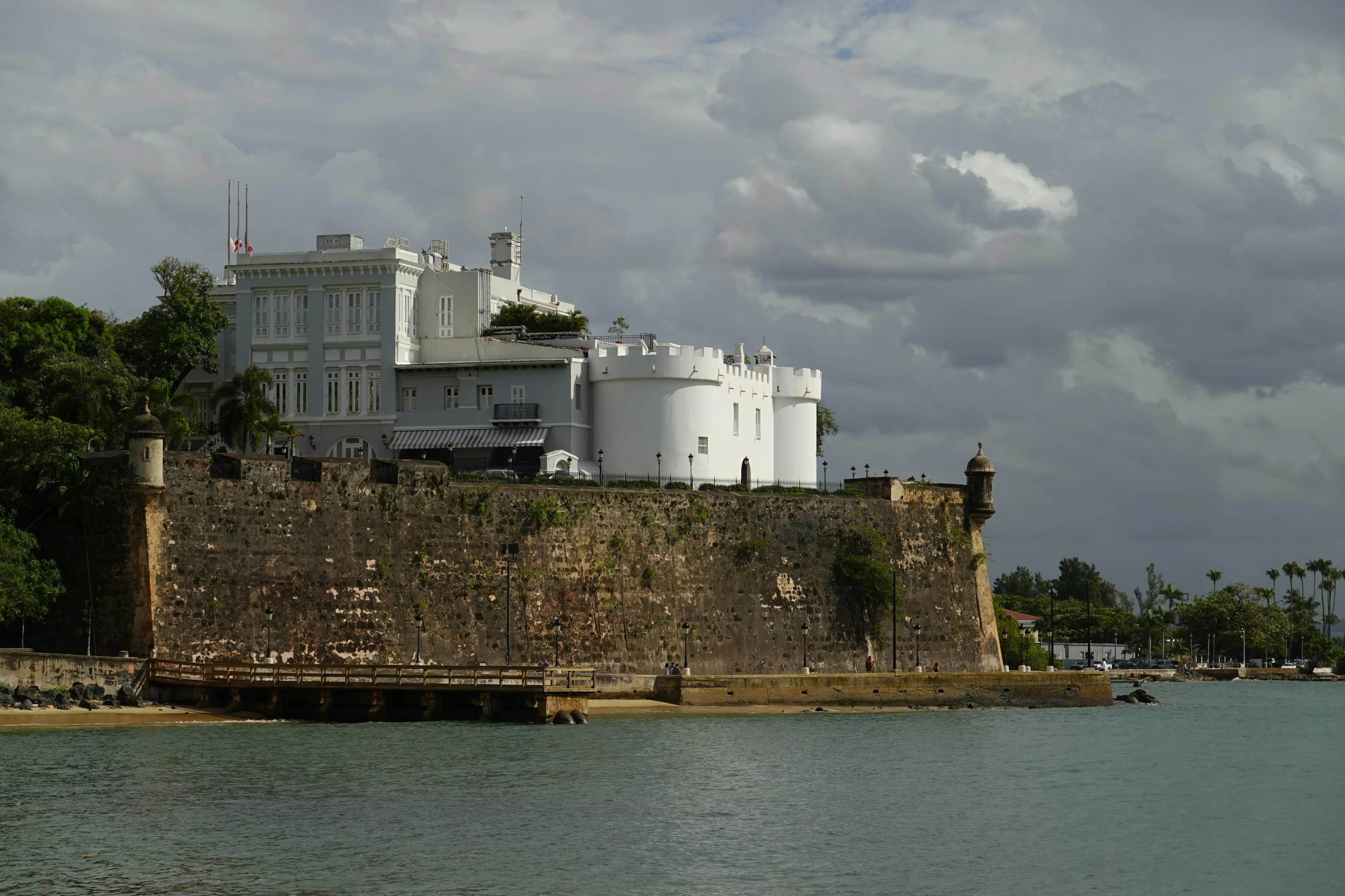 Foto a distancia de la Fortaleza, se observa muro histórico de piedras y el mar con La Fortaleza al tope. Foto por  Richard Lu en Unsplash.