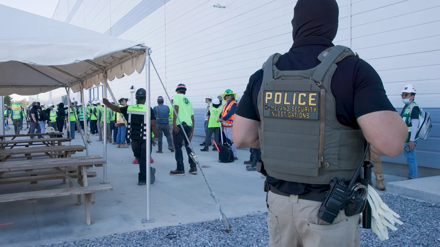 Agente de Homeland Security Investigations de ICE vigila a decenas de trabajadores con cascos y chalecos en fila frente a una planta en Savannah, Georgia, durante un operativo laboral. Imagen suministrada.