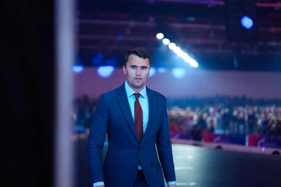 Charlie Kirk, líder conservador estadounidense y fundador de Turning Point USA, aparece en traje frente a un público en un auditorio iluminado.
