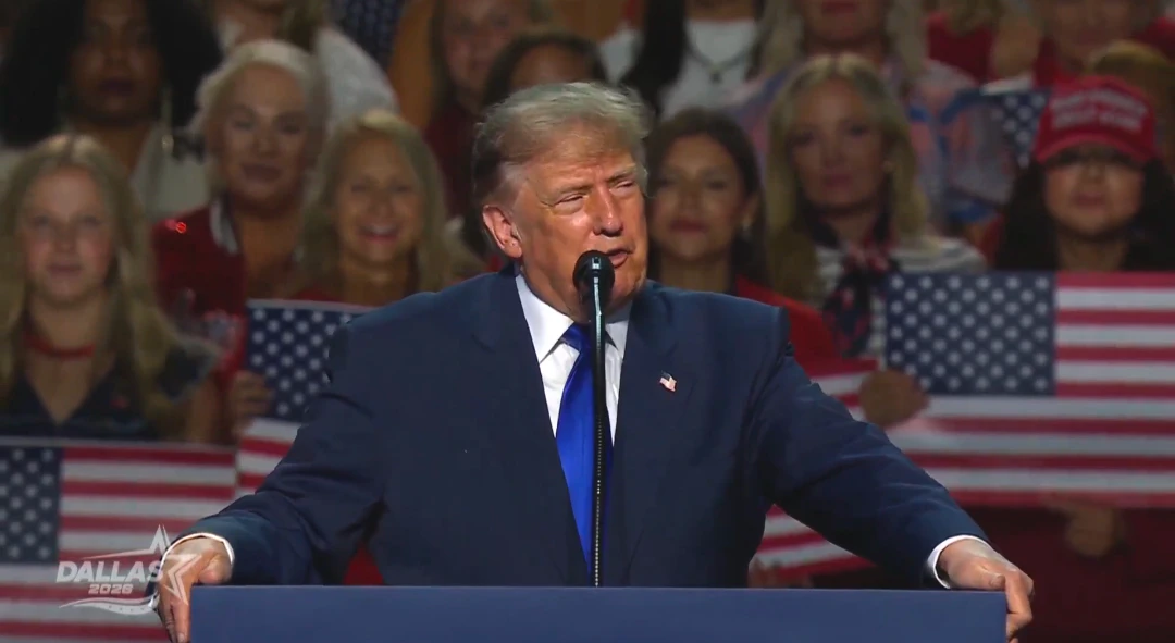 Donald Trump habla frente a un micrófono desde un podio azul oscuro, vestido con traje azul marino, camisa blanca, corbata azul y un pin de la bandera americana en la solapa, con ambas manos apoyadas en los bordes del podio. Detrás, un público en gradas sostiene banderas de Estados Unidos, con una persona con gorra roja a la derecha, y en la esquina inferior izquierda aparece el logo "Dallas 2026" de la transmisión.
