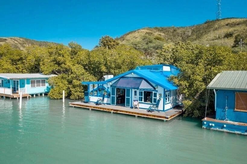 Vista panorámica de una casa flotante azul en La Parguera, Lajas, rodeada de manglares y otras estructuras similares sobre el agua, tomada de un anuncio de alquiler en Join a Join.
