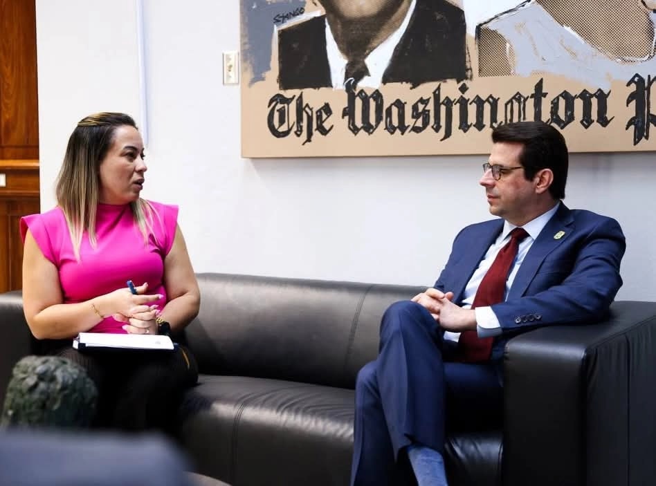 Itza García, wearing a fuchsia sleeveless top and holding a pen and documents, sits on a black leather sofa speaking with Francisco Domenech, who wears glasses, a navy suit and a dark red tie, seated in an armchair facing her. Behind them hangs a large artwork of The Washington Post front page.